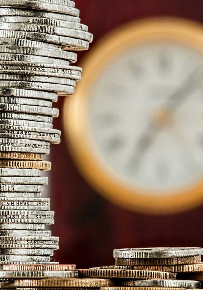 A close-up image of stacked coins with a blurred clock, symbolizing time and money relationship.